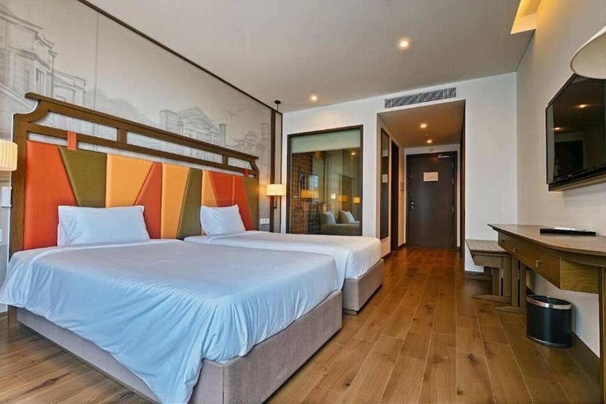 Ein Hotelzimmer mit zwei Betten, Holzboden, Schreibtisch mit Stuhl, großem Spiegel und Fenster mit Blick auf ein Gebäude.