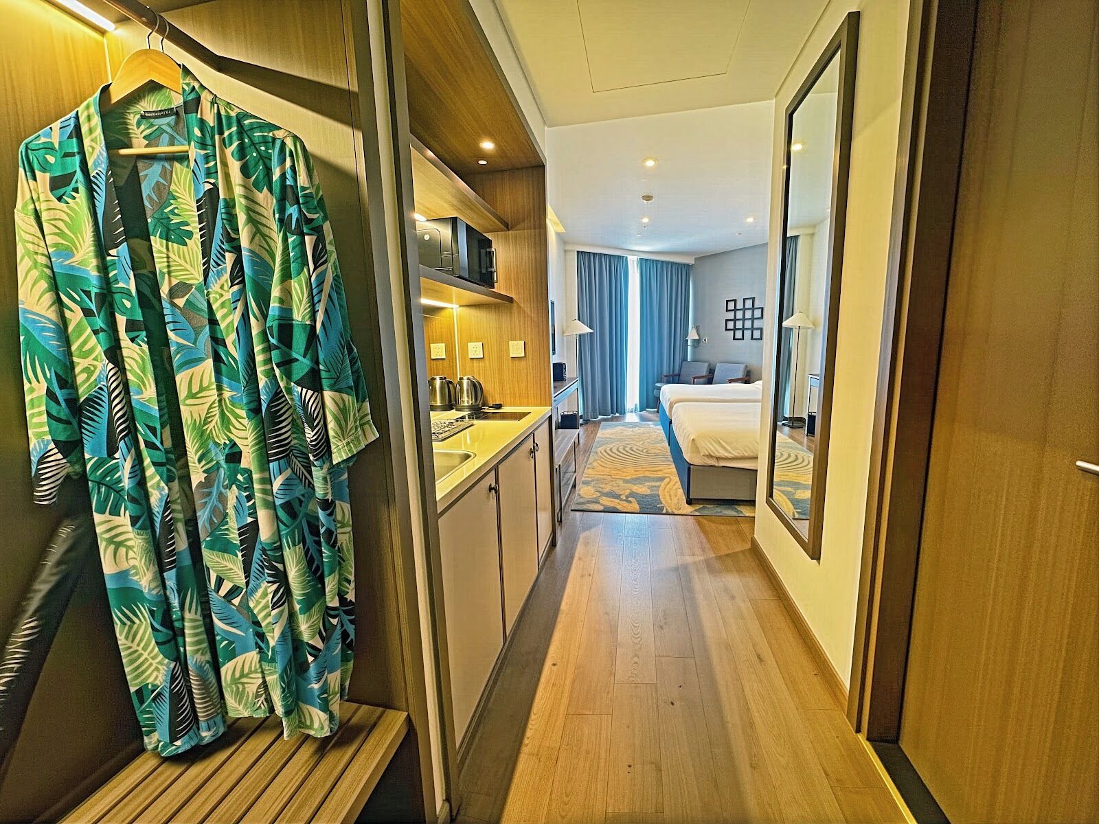 A hotel room with a wooden floor, a bed, a desk with a chair, a mirror, and a bathroom with a shower and toilet.