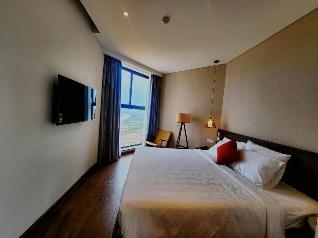 A hotel room with a large bed, a flat-screen TV mounted on the wall, a chair, a lamp, and a view of the outdoors through the window.