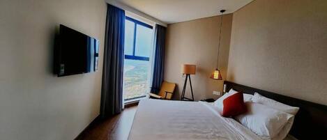 A hotel room with a large bed, a flat-screen TV mounted on the wall, a chair, a lamp, and a view of the outdoors through the window.