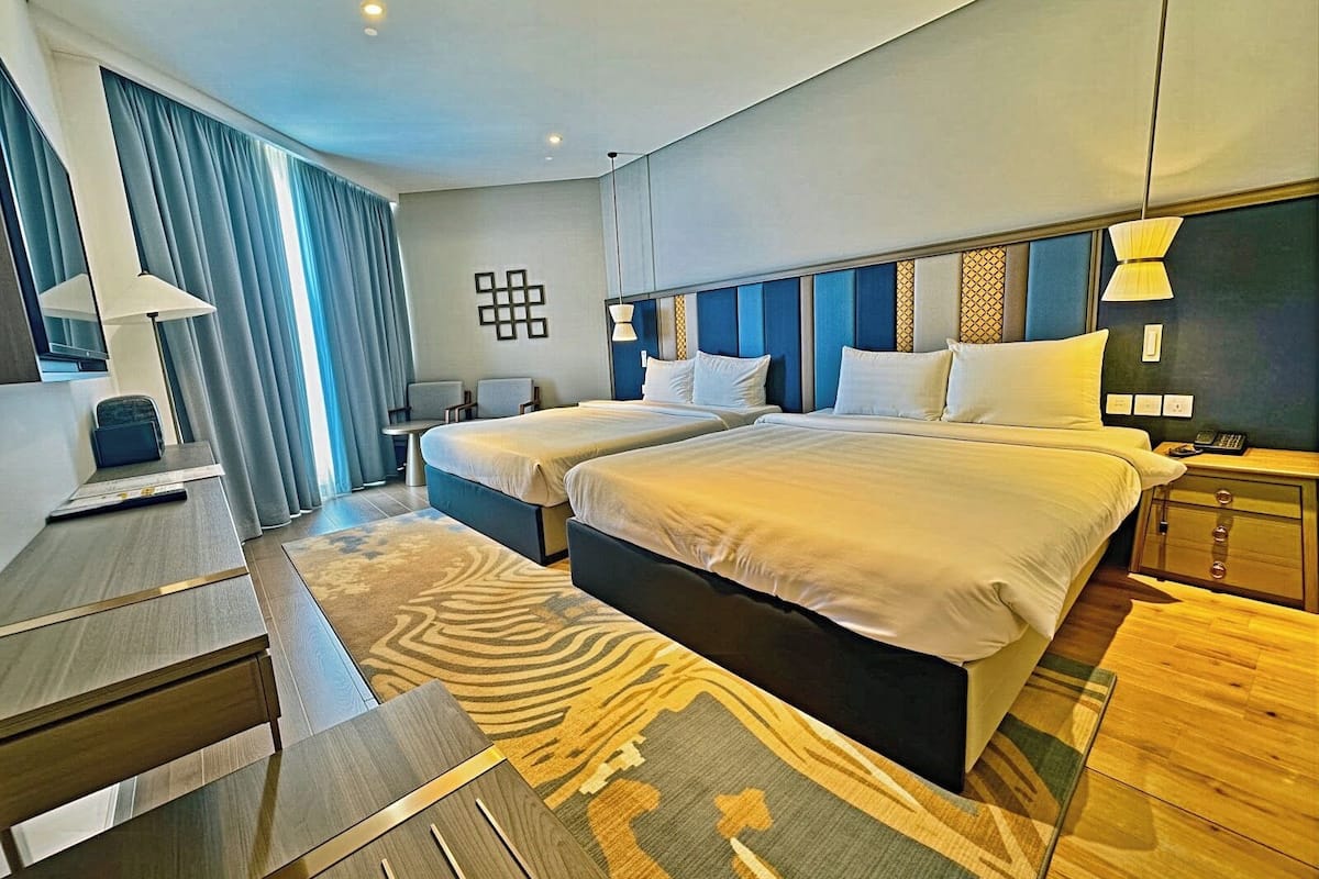Ein Hotelzimmer mit zwei Betten, einem Schreibtisch, einem Stuhl, einer Lampe und einem gemusterten Teppich.