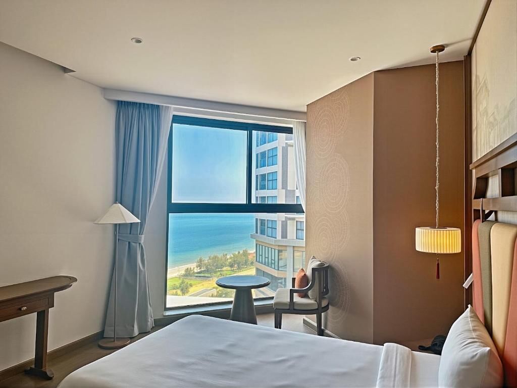A hotel room with a large window offering a view of the sea and buildings, a bed, a small table, and a chair.