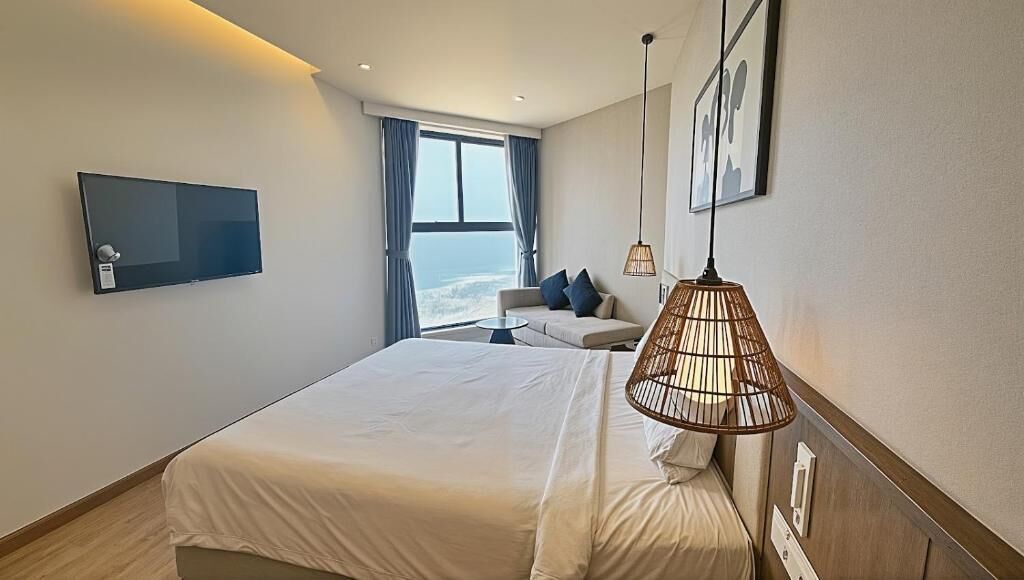 A modern hotel room with a large bed, a sofa, a television, and a view of the sea.