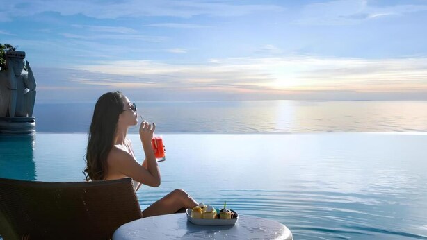 Una persona seduta a bordo piscina con vista sul tramonto, un piatto di cibo e una bevanda.