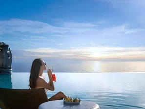 En person sidder ved en pool med udsigt til solnedgang, en tallerken mad og en drik.