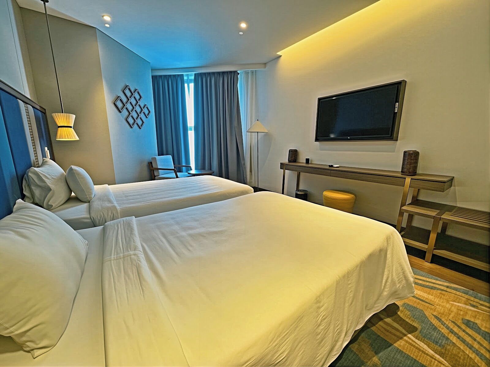 A hotel room with two beds, a desk, a TV, and a window with curtains.