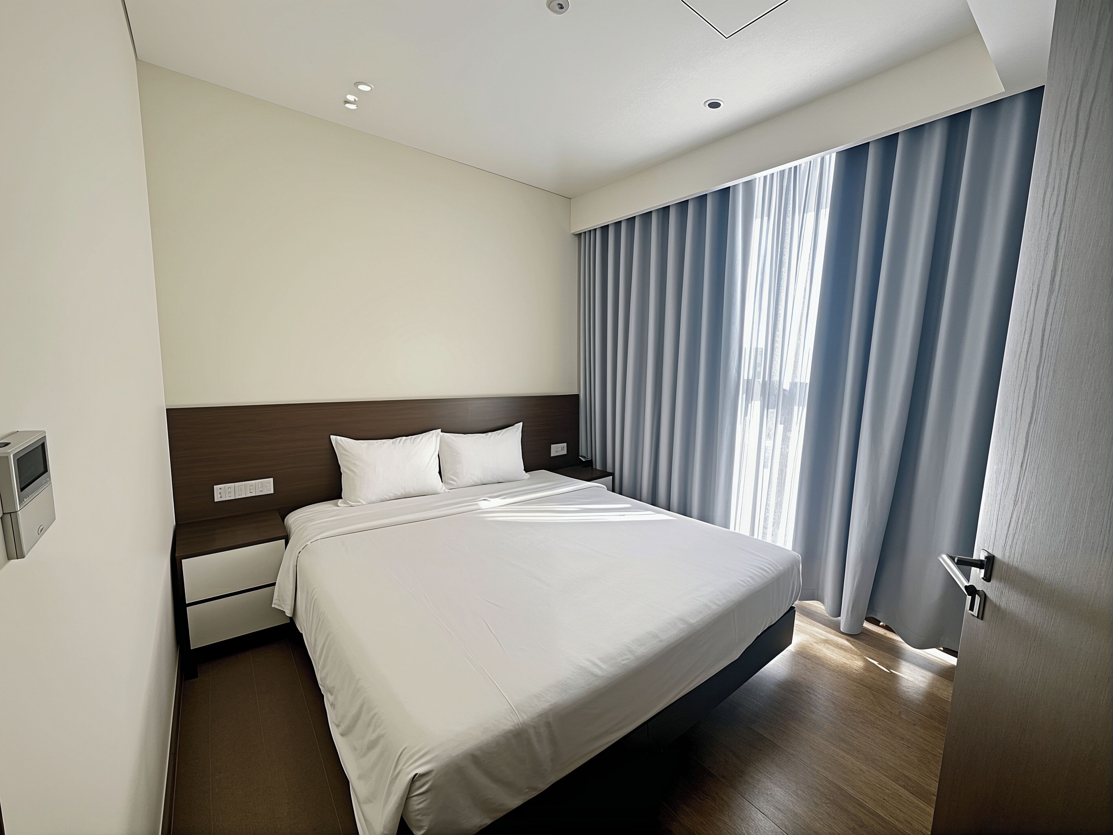 A modern hotel room with a large bed, a wooden headboard, a bedside table, and a window with curtains.