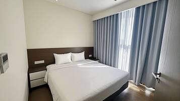A modern hotel room with a large bed, a wooden headboard, a bedside table, and a window with curtains.