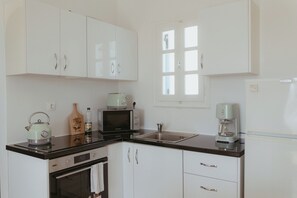 A kitchen with white cabinets, a black countertop, a microwave, a kettle, a coffee maker, and a window with blinds. - Sunset Getaway Aliki: Charming apartment by the beach with beautiful sunset view (Aliki)