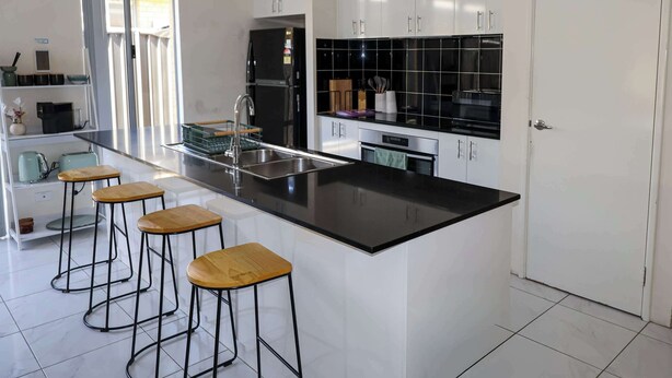 Cozinha moderna com armários brancos, bancada preta e três banquetas de madeira.