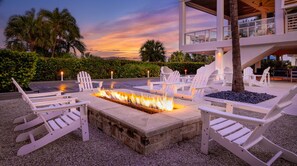 Un foyer entouré de chaises Adirondack blanches, avec en arrière-plan un bâtiment moderne et des palmiers, le tout baigné par les couleurs du coucher de soleil.