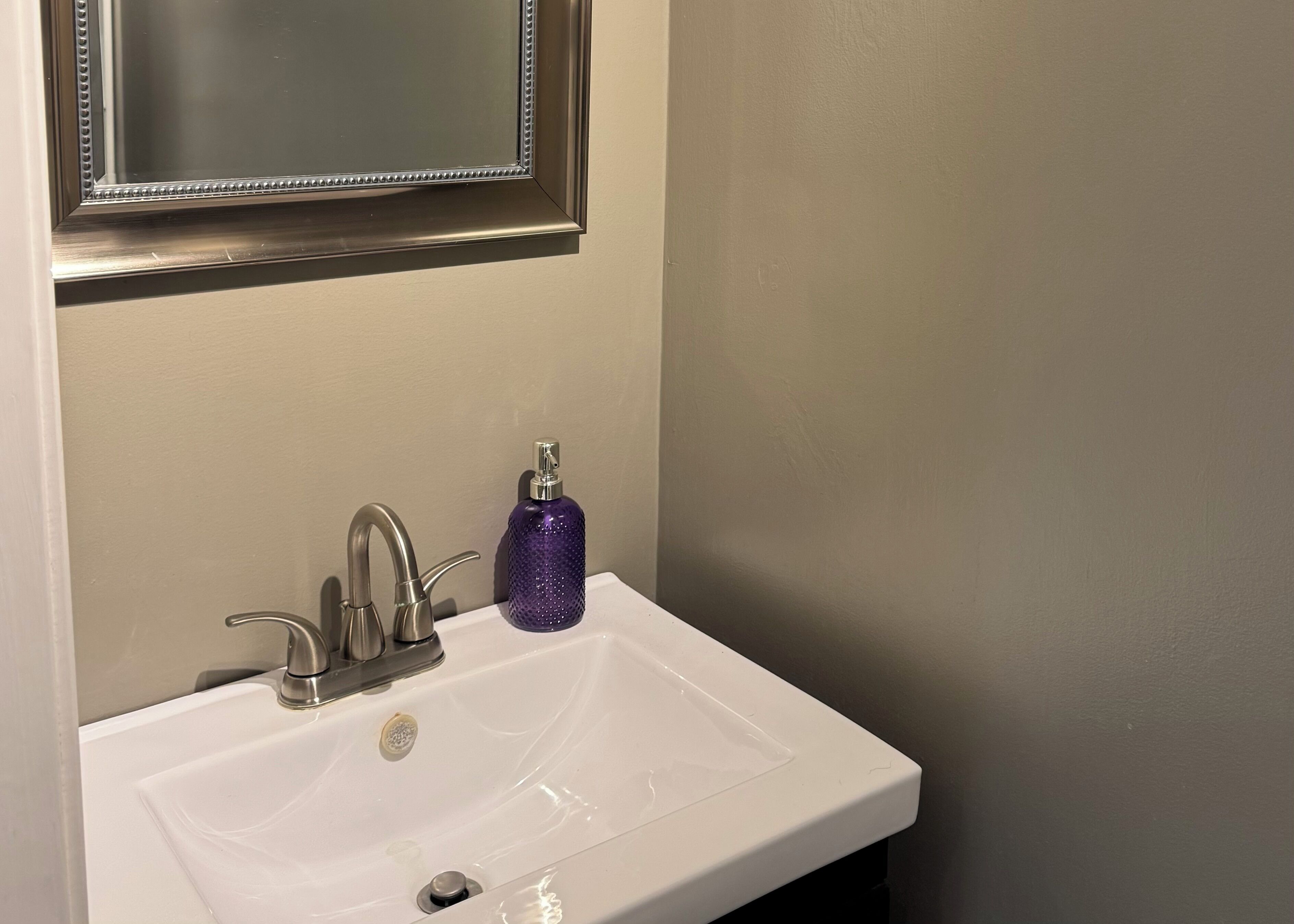 Ein Waschbecken mit Spiegel darüber, einem Wasserhahn und einem violetten Seifenspender.