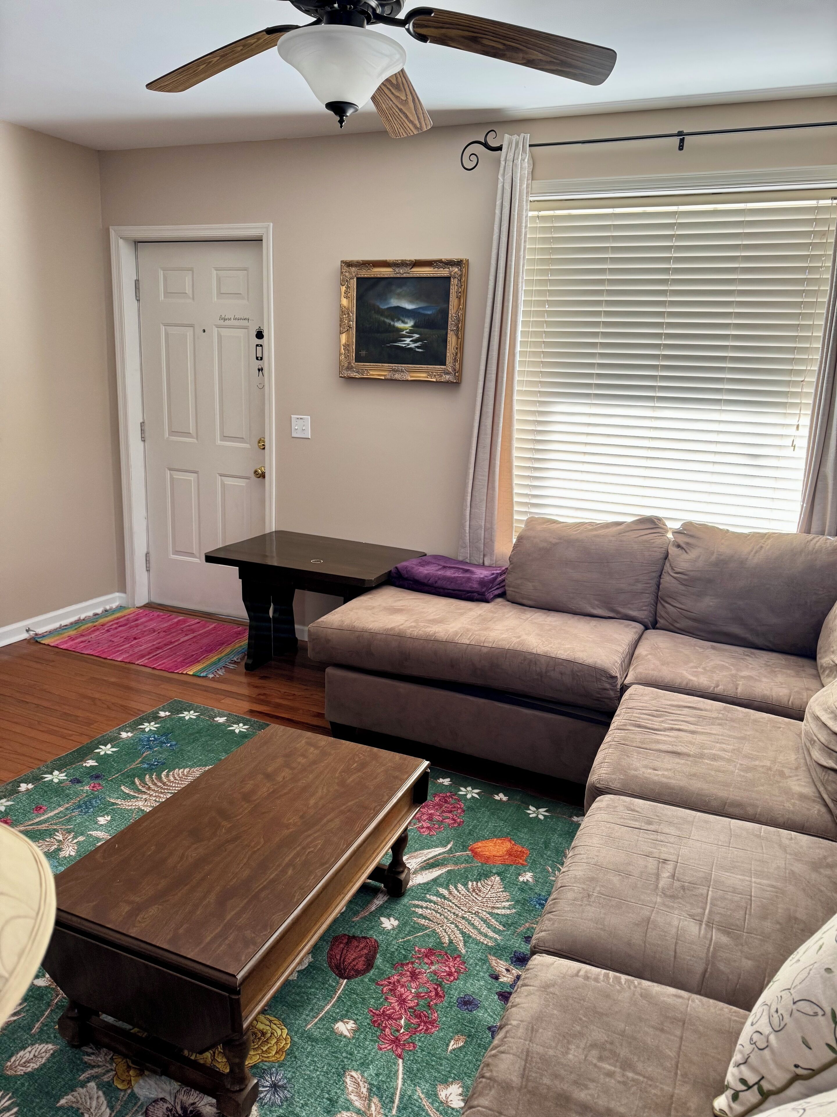 A living room with a beige sofa, a wooden coffee table, a ceiling fan, a framed picture on the wall, and a window with blinds.