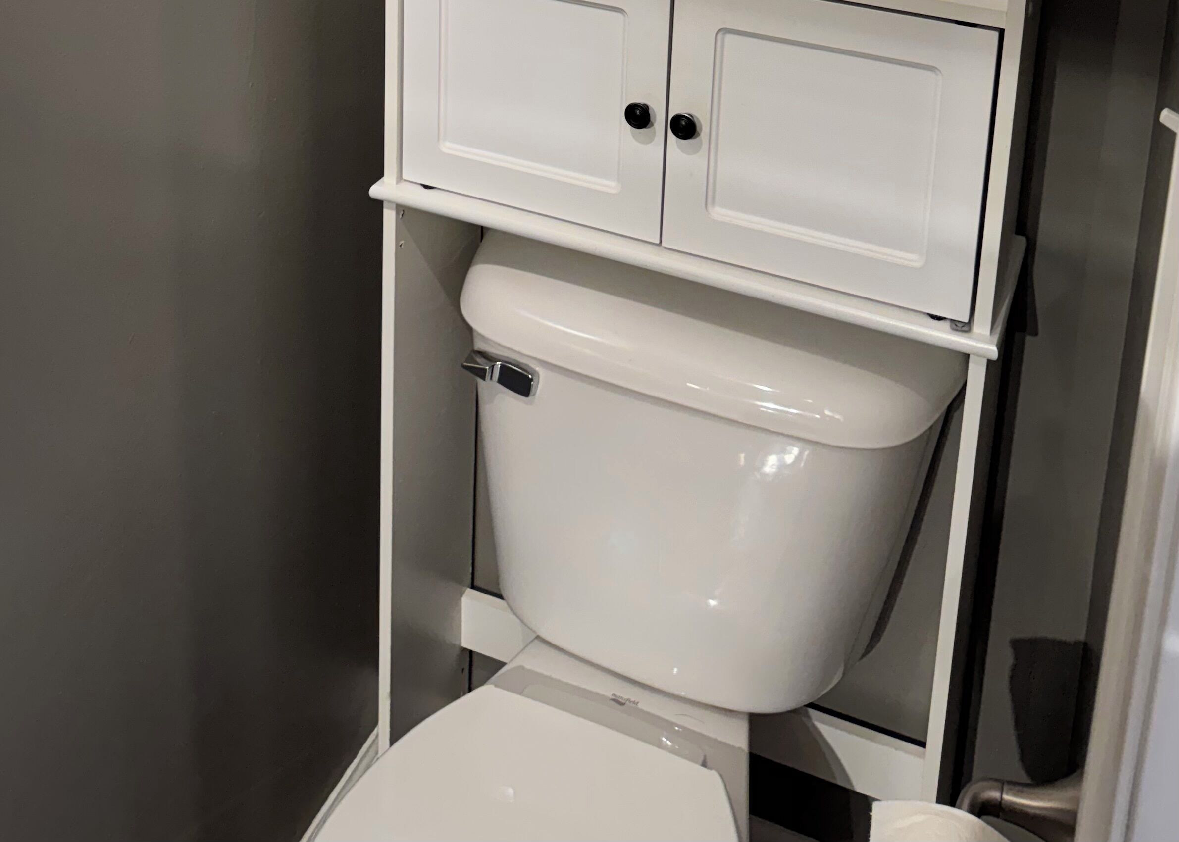 A toilet with a white tank and lid, a chrome flush handle, and a white cabinet above it.