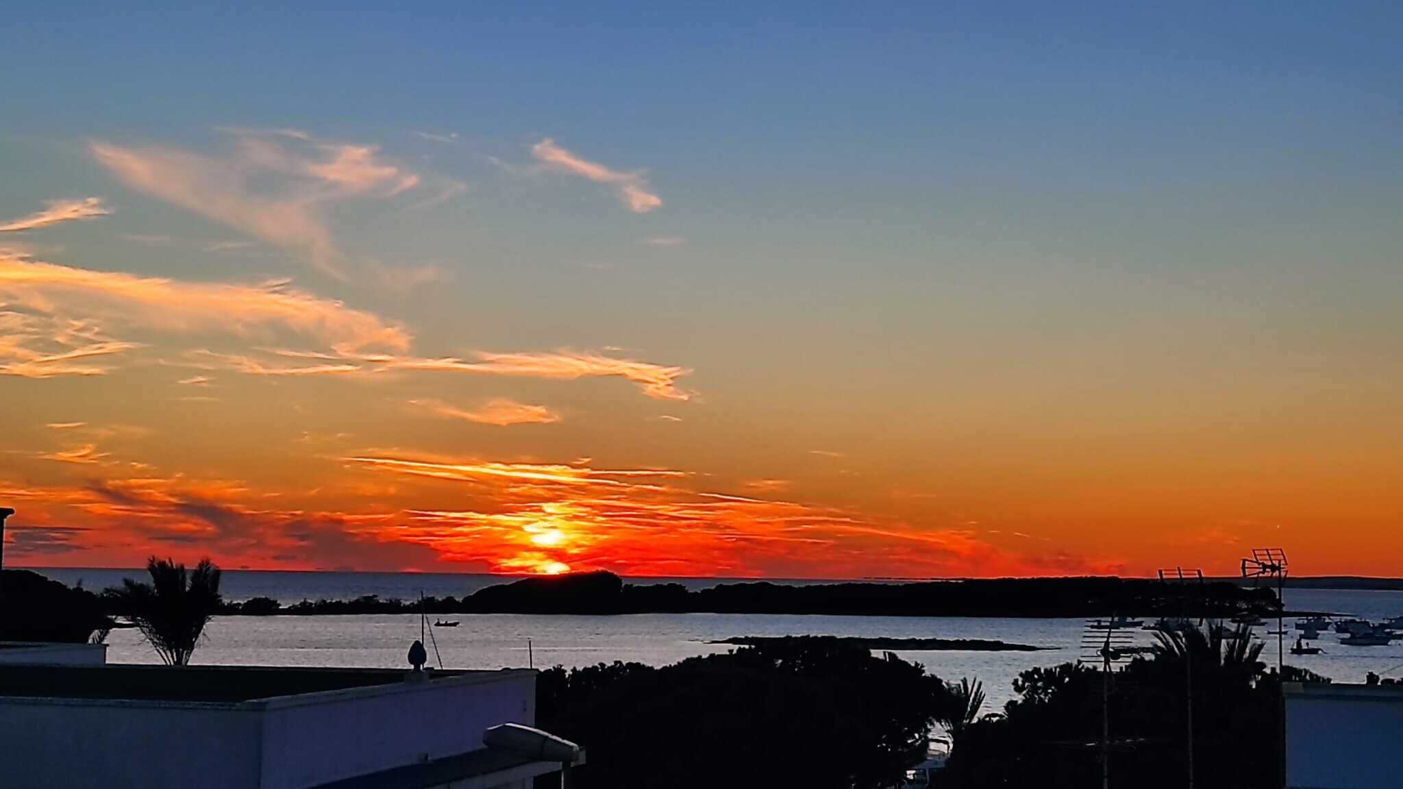 A sunset over a coastal area with silhouettes of buildings and boats.