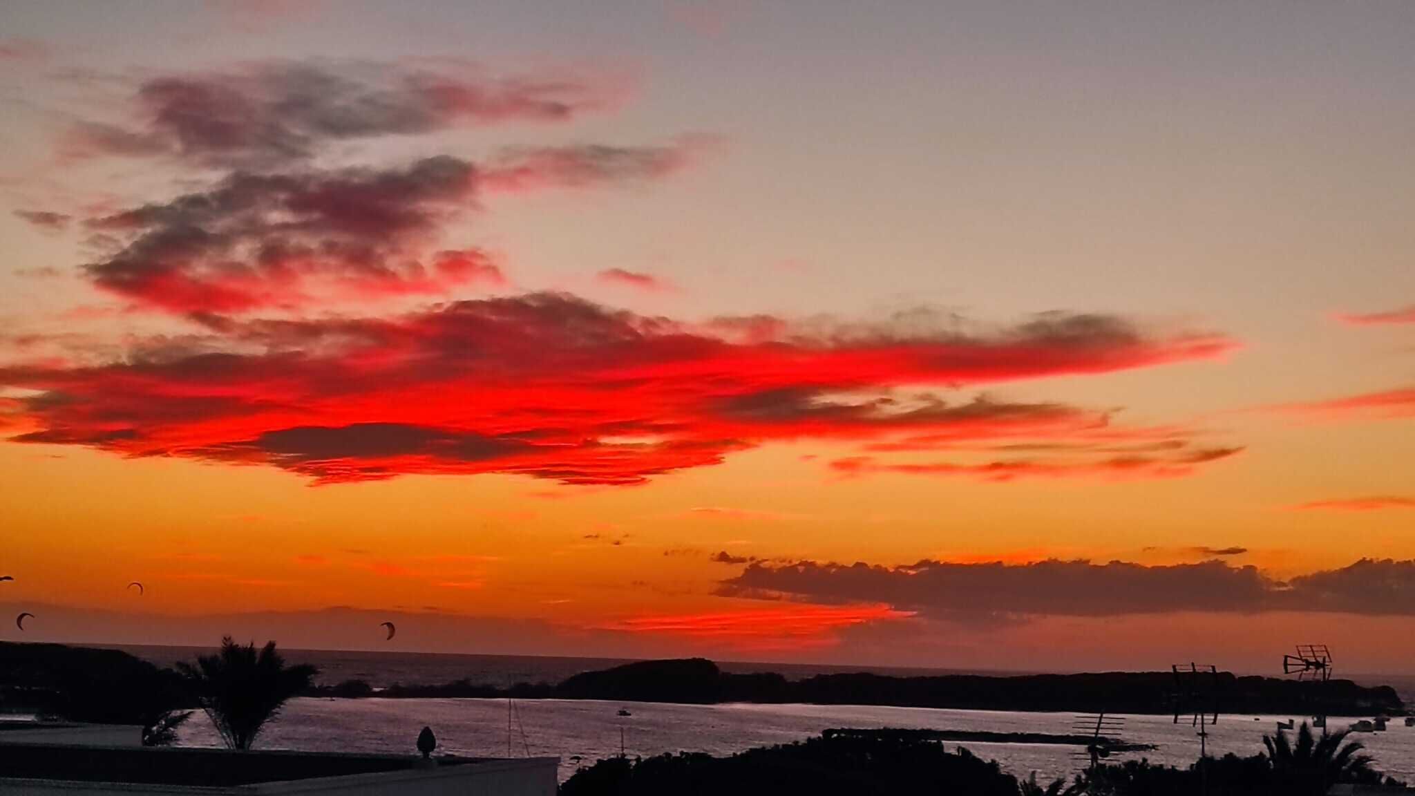 Atardecer con nubes rojas intensas sobre un cuerpo de agua, siluetas de árboles y formas de terreno a lo lejos.