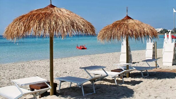 Een strand met rieten parasols, witte lounge stoelen en een heldere blauwe zee.