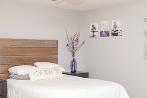Une chambre à coucher avec une tête de lit en bois, un lit recouvert de linge de lit blanc, une table de chevet ornée d’un vase violet et trois tableaux encadrés accrochés au mur.