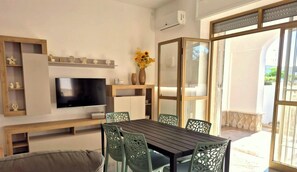 A modern living room with a flat-screen TV, wooden shelves, a dining table with chairs, and a large glass door leading outside. - Villa Albachiara with veranda (Torre Lapillo)