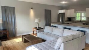 Una sala de estar moderna con un sofá, una mesa de centro y una pequeña mesita auxiliar. Hay una zona de cocina con un refrigerador y armarios.