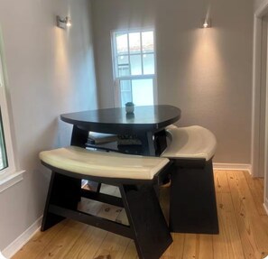 A small dining area with a round table and bench, a window, and a light fixture on the wall.