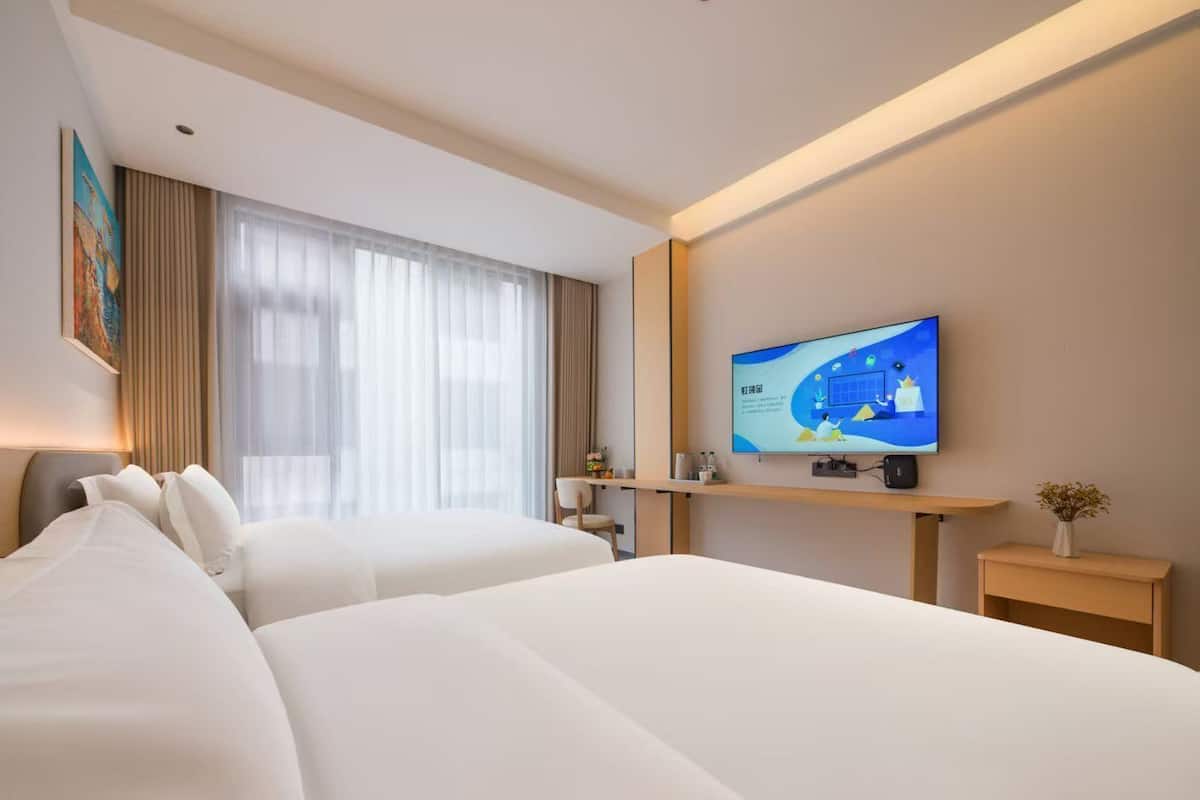 A hotel room with two beds, a TV on the wall, a desk, and a window with curtains.