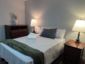 Ein ordentlich bezogenes Bett mit einer gemusterten Tagesdecke, zwei Kissen und einer grünen Zudecke, sowie ein Nachttisch mit einer Lampe.