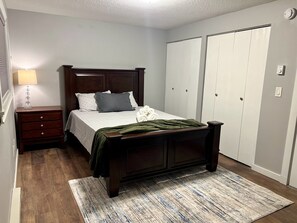 Quarto com cama de madeira, mesa de cabeceira, abajur, manta verde e guarda-roupa com portas deslizantes.