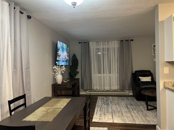 A living room with a TV, a dining table with chairs, a sofa, and a large plant.