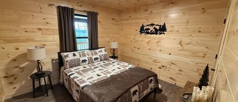 Un dormitorio de cabaña de madera con una cama, dos mesitas de noche, una lámpara, una silla y una pequeña mesa con velas.