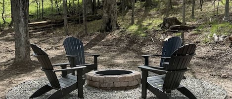 Eine runde Sitzgruppe mit einem Feuerkorb in der Mitte, umgeben von vier Adirondack-Stühlen, gelegen in einem bewaldeten Gebiet.