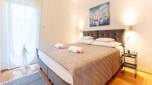 Un letto rifatto con precisione, con copriletto e cuscini color beige, un quadro con velieri sulla parete e un comodino con una lampada.