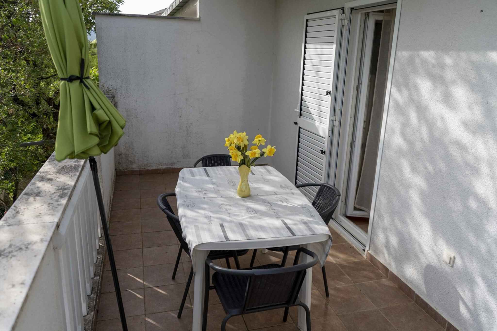 Een klein terras met een tafel en stoelen, een gele vaas met bloemen en een groene parasol.