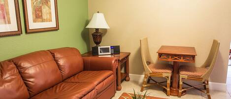 A living room with a brown leather sofa, a wooden table with two wicker chairs, a lamp, and a small potted plant.