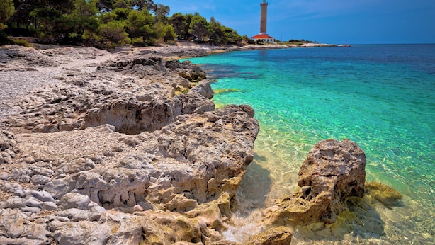Un paisaje costero con terreno rocoso, aguas turquesas cristalinas y un faro a lo lejos.