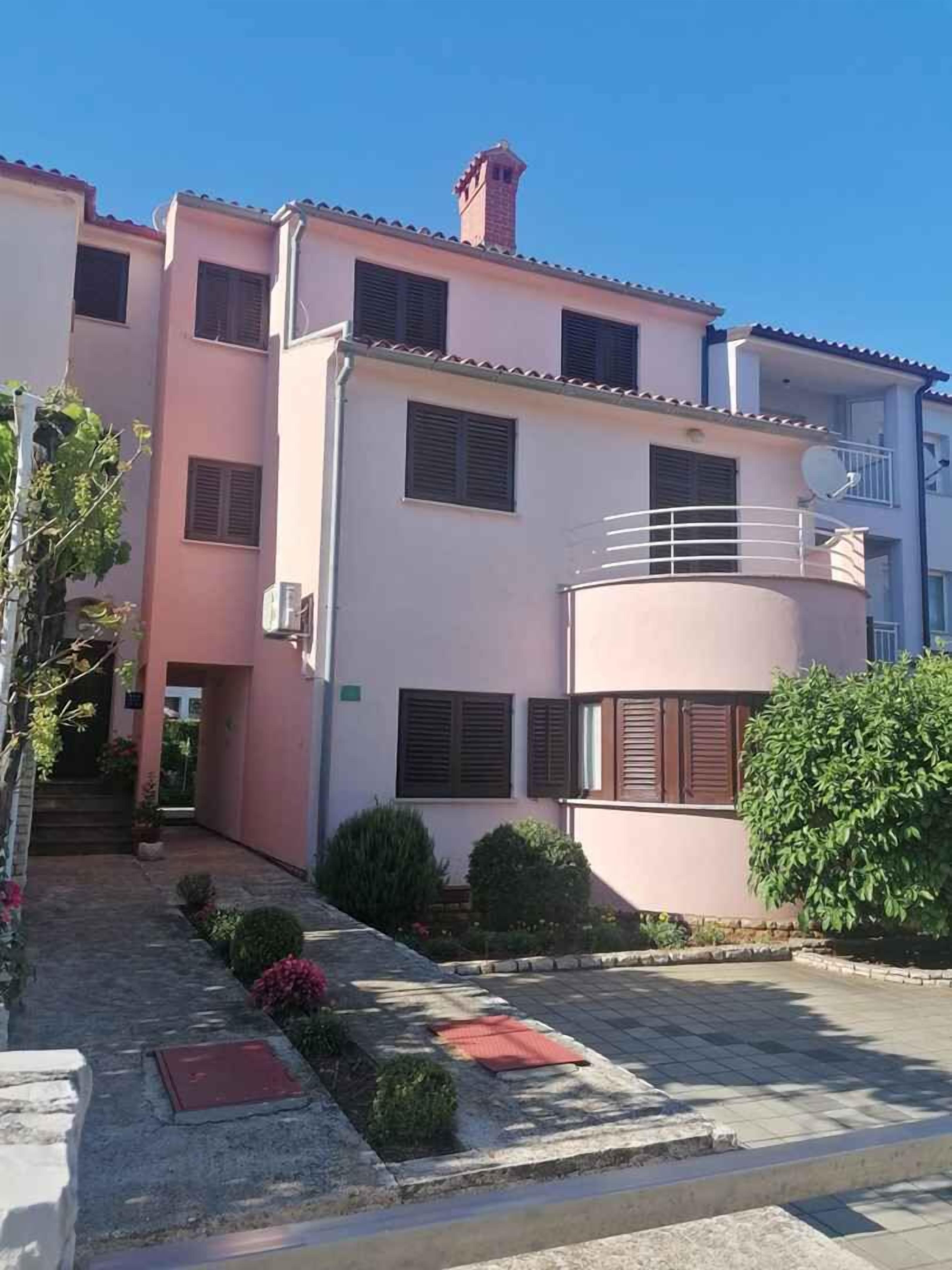 Un immeuble résidentiel à plusieurs étages, doté d’une façade rose, de nombreuses fenêtres avec volets et d’un petit balcon.