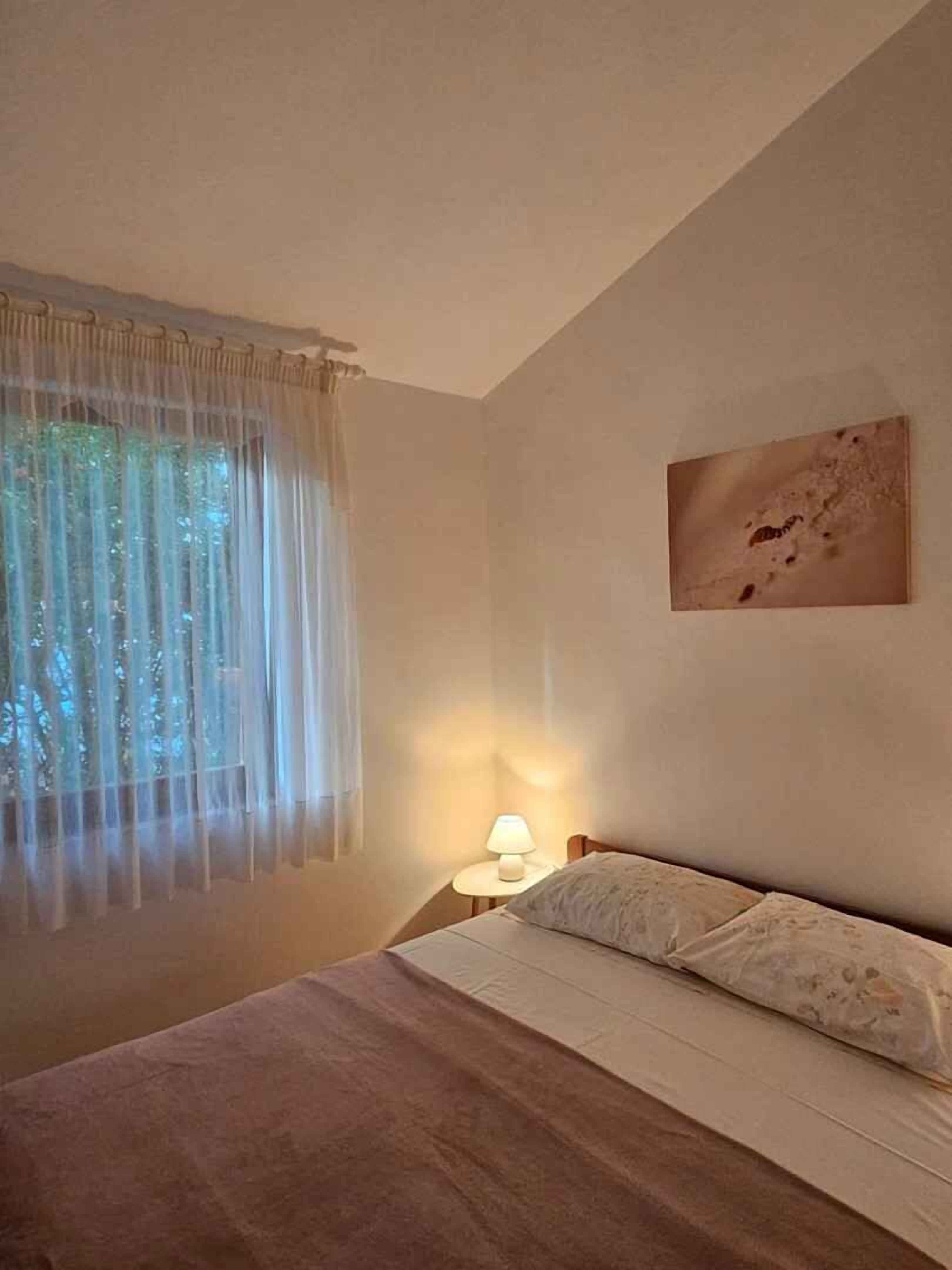 A bedroom with a bed, a bedside lamp, a window with sheer curtains, and a wall-mounted picture.