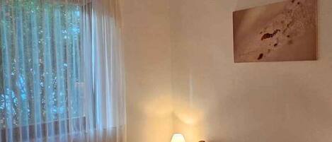 A bedroom with a bed, a bedside lamp, a window with sheer curtains, and a wall-mounted picture.