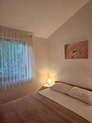 Ein Schlafzimmer mit Bett, Nachttischlampe, einem Fenster mit durchsichtigen Vorhängen und einem an der Wand hängenden Bild.
