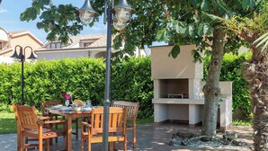 Zona pranzo all'aperto con tavolo e sedie in legno, circondata dal verde e dotata di un barbecue.