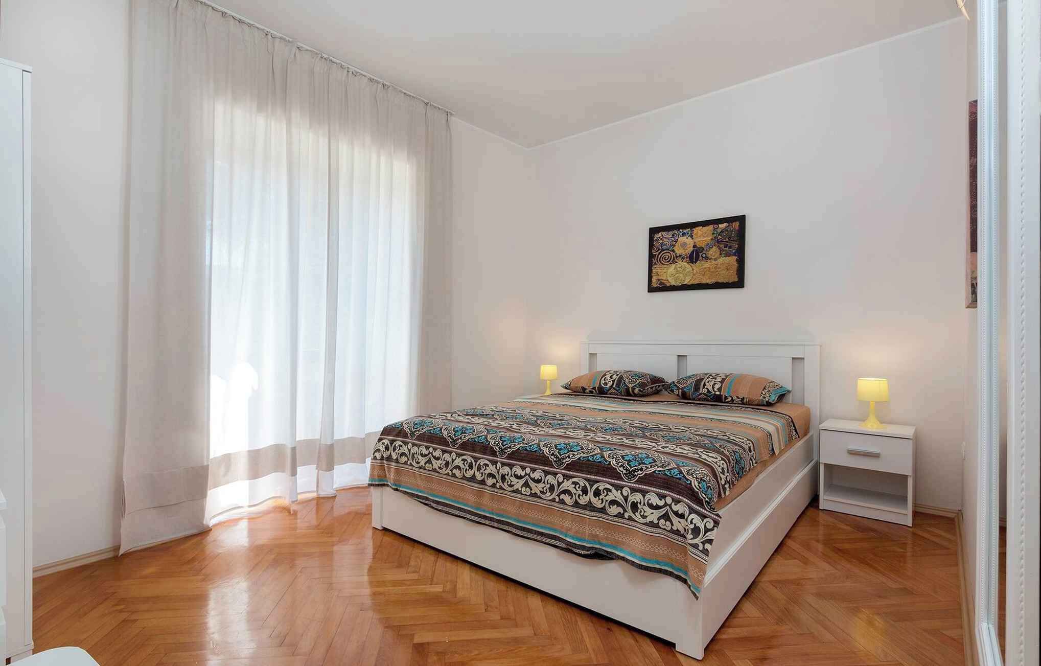A bedroom with a large bed, two bedside tables with lamps, a window with sheer curtains, and a wall-mounted artwork.