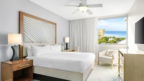 Habitación de hotel con cama, mesitas de noche, escritorio, silla, ventilador de techo, ventana grande con vista al océano y televisión.