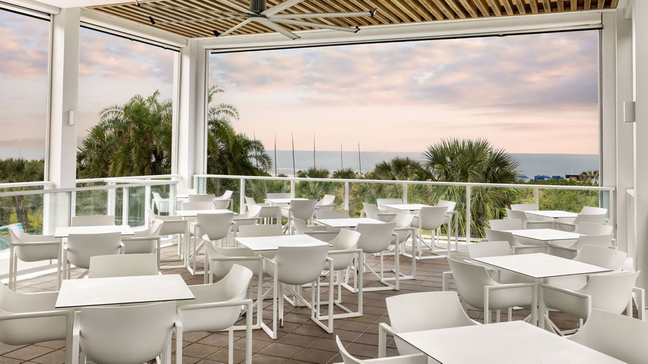 A terrace with white tables and chairs, large windows, and a view of the ocean.