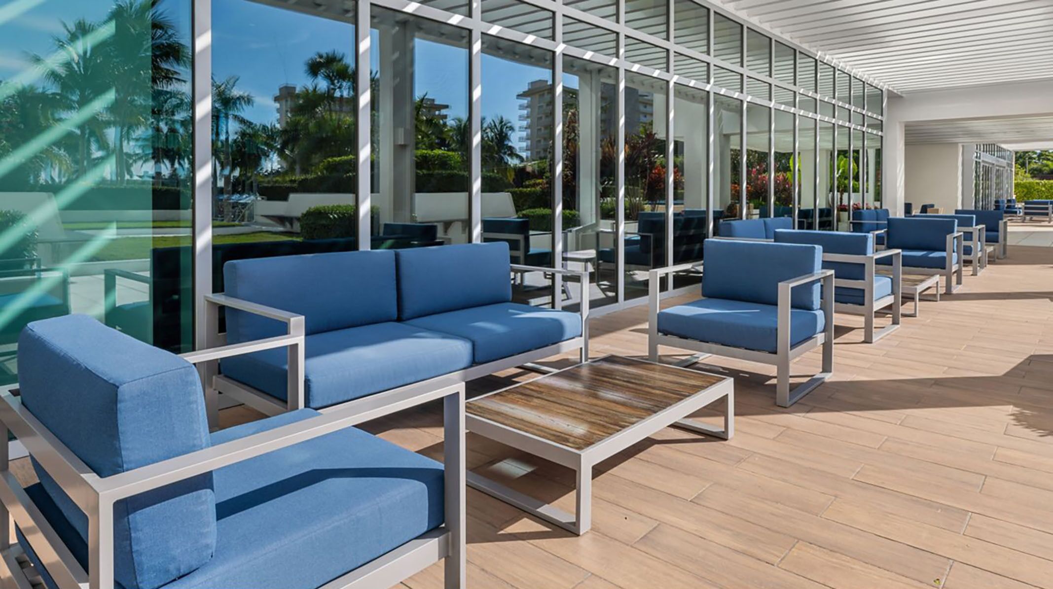 Un espace extérieur moderne, aménagé avec des chaises et des tables bleues dotées de coussins, de grandes fenêtres en verre et une terrasse en bois.