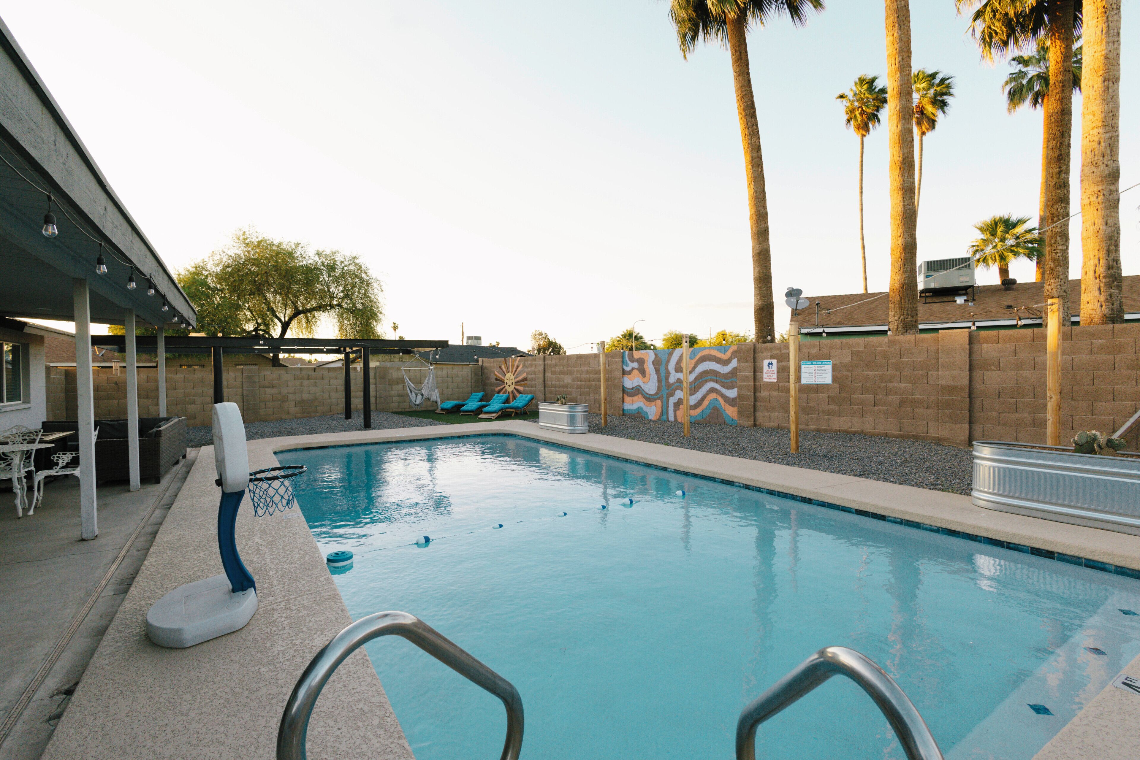 A swimming pool with a basketball hoop, lounge chairs, palm trees, and a covered seating area.