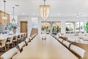 Un vaste espace dédié à la salle à manger, avec une longue table en bois et de nombreuses chaises, un lustre moderne et une vue sur l’extérieur grâce à de grandes fenêtres.