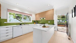 A modern kitchen with white cabinetry, a central island, and a view of the garden through the window. - Elegant 4BR Sanctuary with Private Pool (Warrawee)
