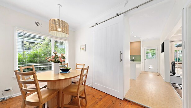 A modern dining area with a wooden table and chairs, a potted plant, and a sliding door leading to a kitchen. - Elegant 4BR Sanctuary with Private Pool (Warrawee)