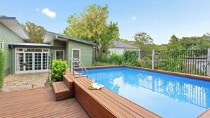A swimming pool with wooden deck seating and a glass door leading to a house. - Elegant 4BR Sanctuary with Private Pool (Warrawee)