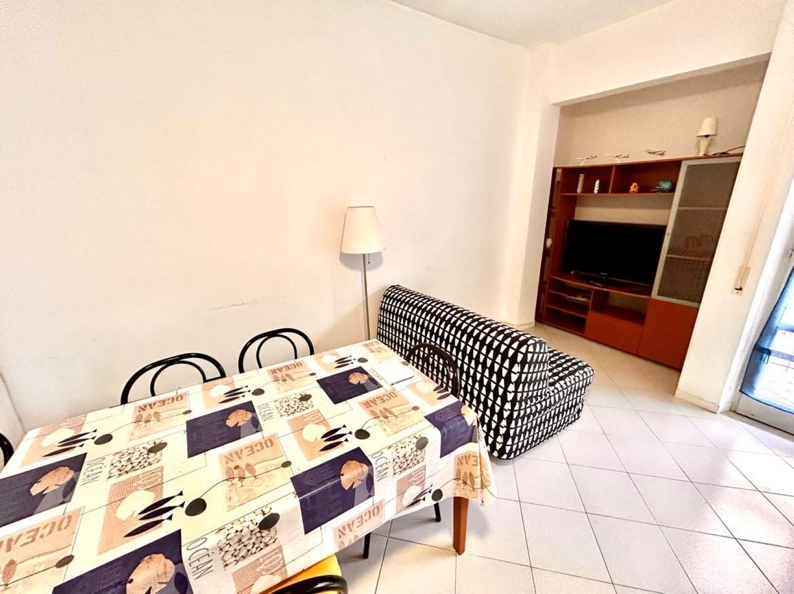 A dining area with a table covered in a patterned tablecloth, a black and white sofa, a lamp, and a TV in the background.